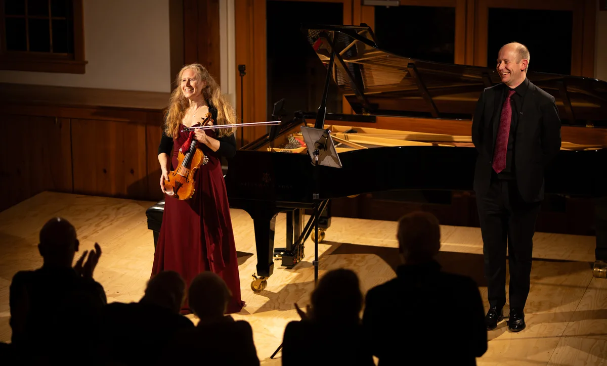 A smiling woman holding a violin stands next to a grand piano with a man in formal attire on stage. Audience members in the foreground applaud under warm lighting in a wood-paneled room.
