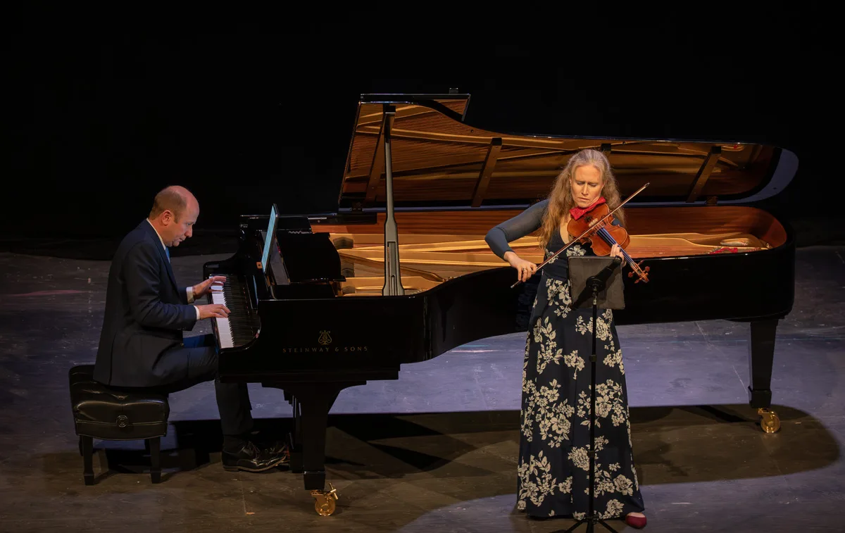 A man in a suit plays a grand piano while a woman in a long floral dress plays the violin beside him on a dimly lit stage.