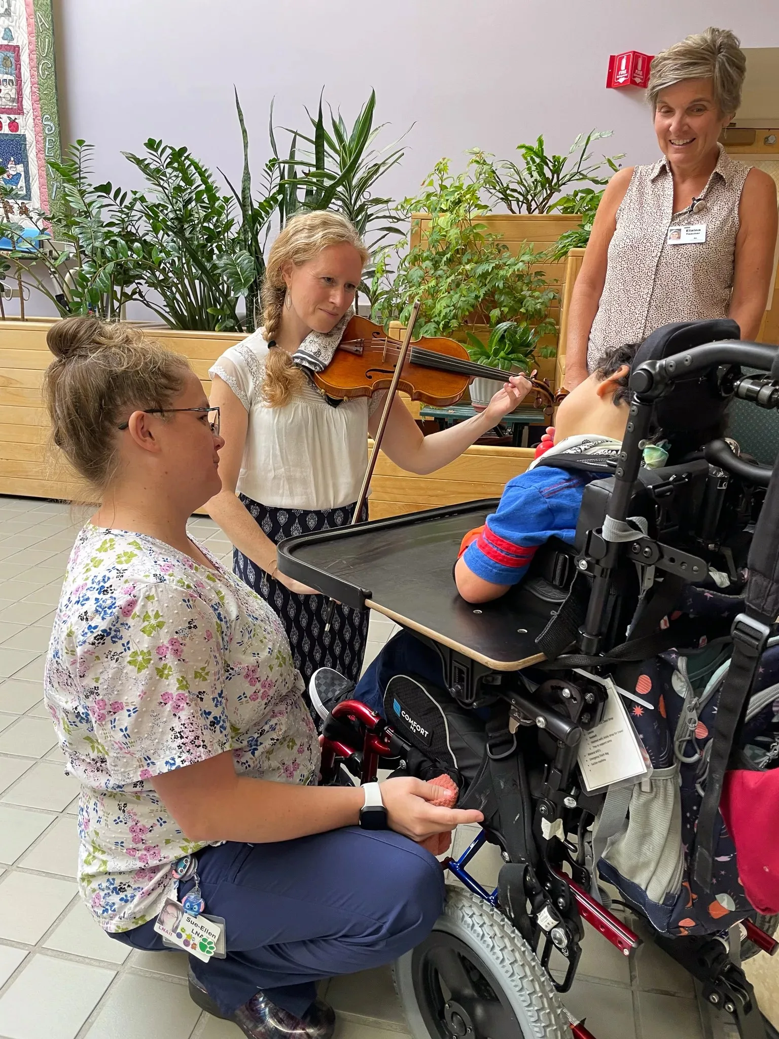 A young woman plays violin for a child in a wheelchair, while two women—one crouching and one standing—smile and watch in a bright indoor space with green plants in the background.