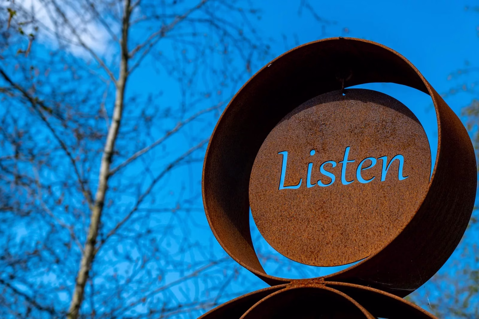 A close-up of a rust-colored metal sculpture with the word Listen cut out, set against a bright blue sky and tree branches in the background.