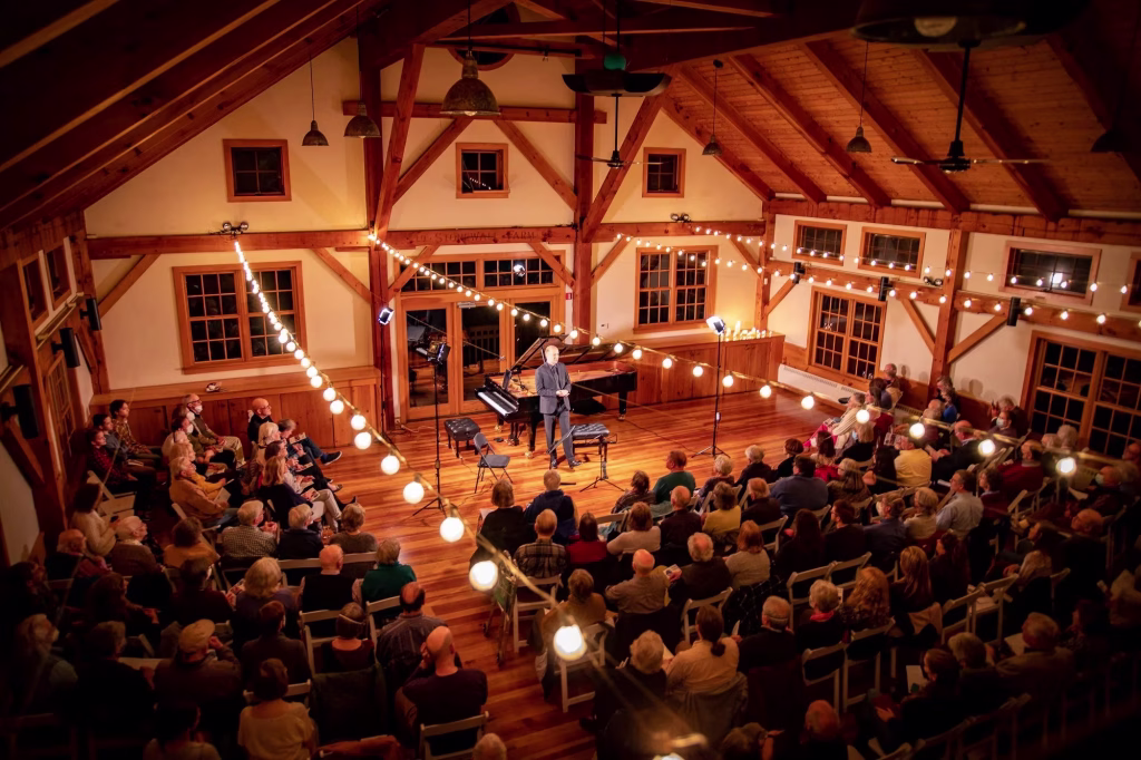 A musician stands on a warmly lit wooden stage in a cozy, rustic hall, surrounded by string lights and an attentive seated audience. A grand piano is onstage, and the atmosphere is inviting and intimate.