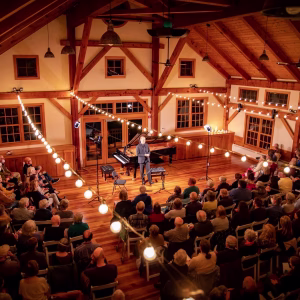 A musician stands on a warmly lit wooden stage in a cozy, rustic hall, surrounded by string lights and an attentive seated audience. A grand piano is onstage, and the atmosphere is inviting and intimate.