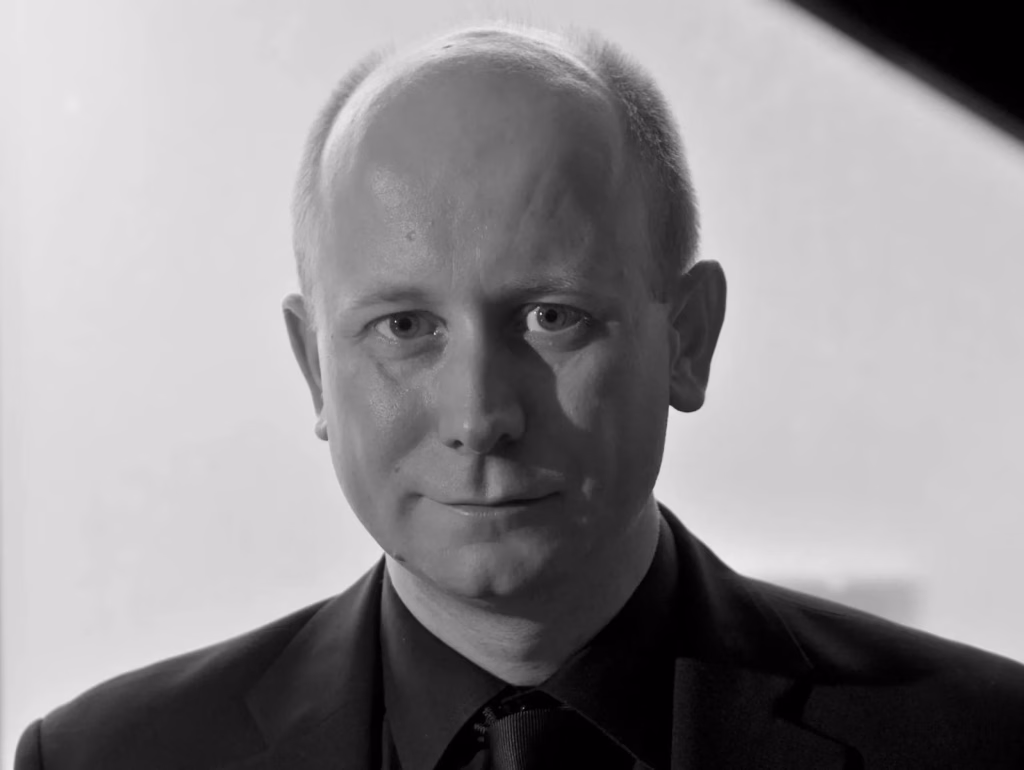 A man with a bald head and light hair, wearing a dark suit and tie, looks directly at the camera with a neutral expression in a black and white portrait.
