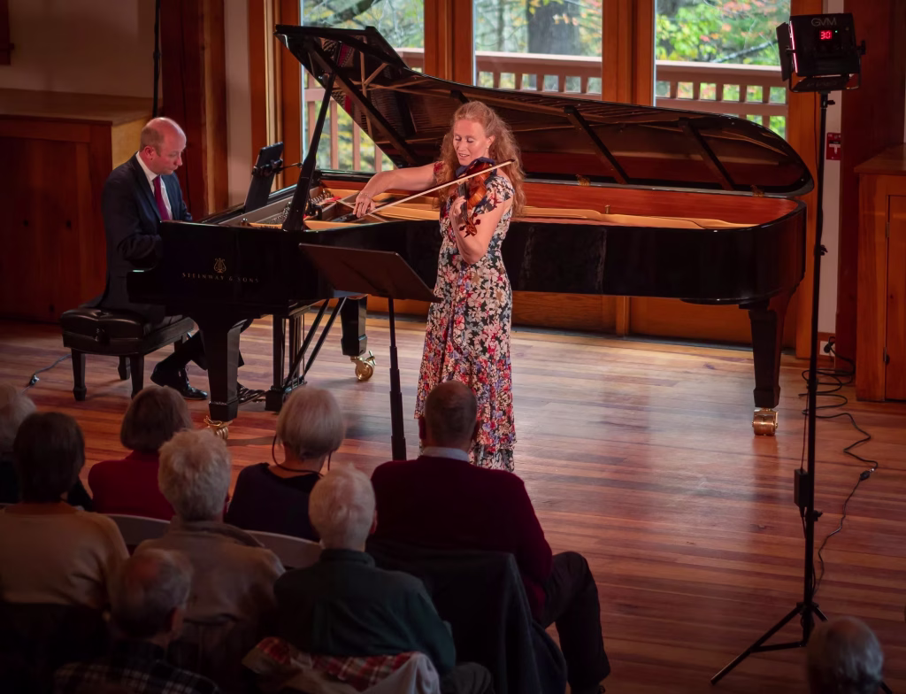A woman in a floral dress plays the violin next to a grand piano, with a man playing the piano. They perform on a wooden stage for an attentive seated audience in a well-lit room with large windows.