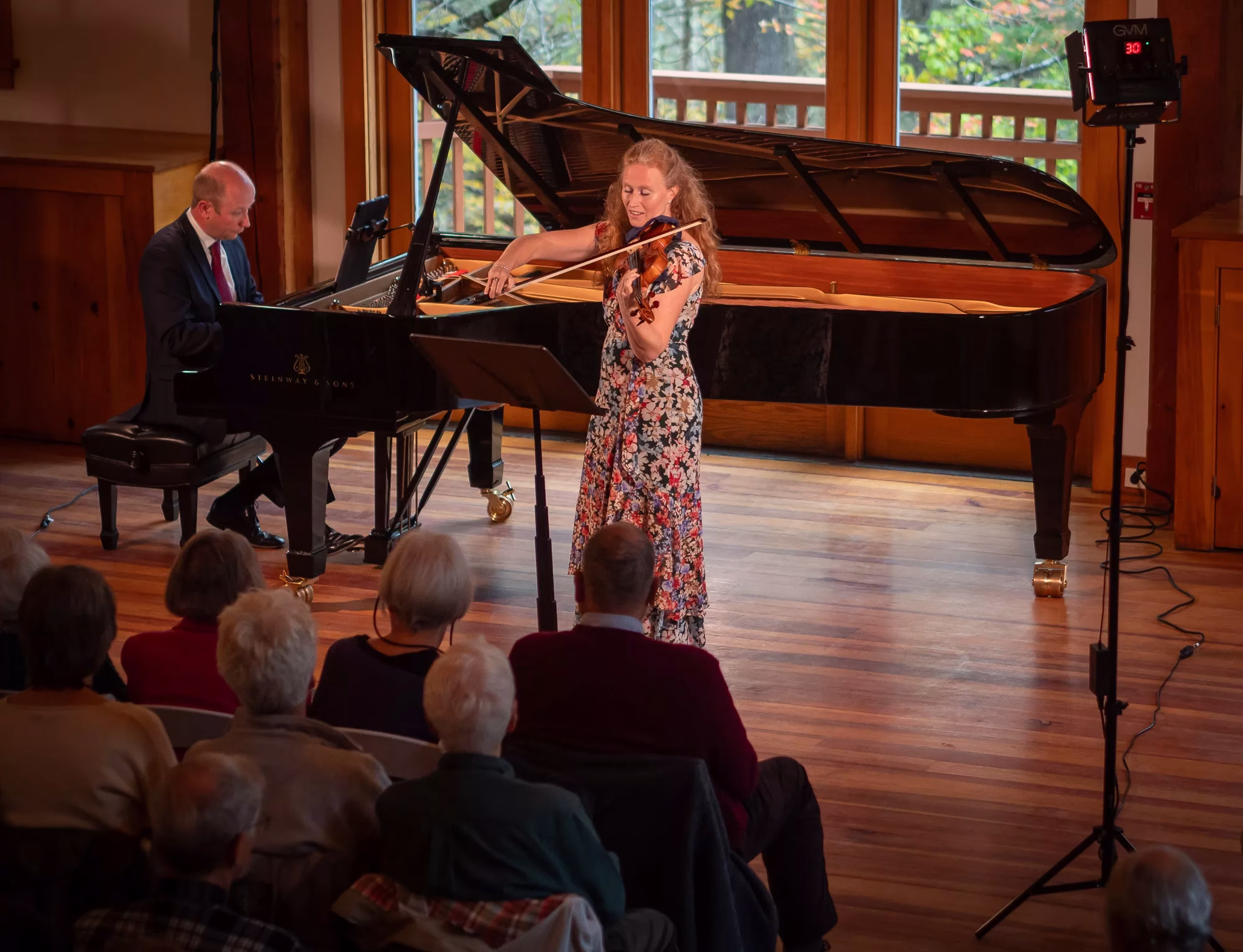 A woman in a floral dress plays the violin next to a grand piano, with a man playing the piano. They perform on a wooden stage for an attentive seated audience in a well-lit room with large windows.