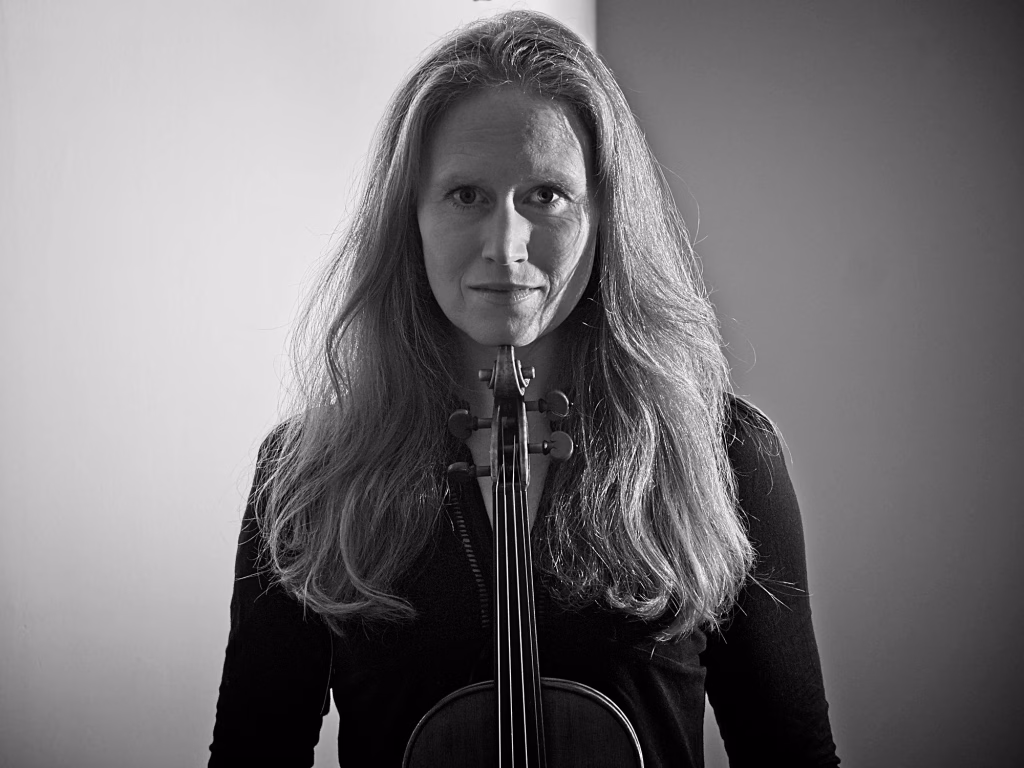 A woman with long hair stands facing the camera, holding a violin upright in front of her. The photo is black and white, with contrasting light and shadow in the background.