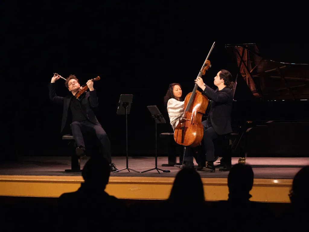Three musicians perform on stage: a violinist dramatically playing, a cellist in mid-performance, and a pianist at a grand piano. The audience watches in silhouette from the darkened foreground.