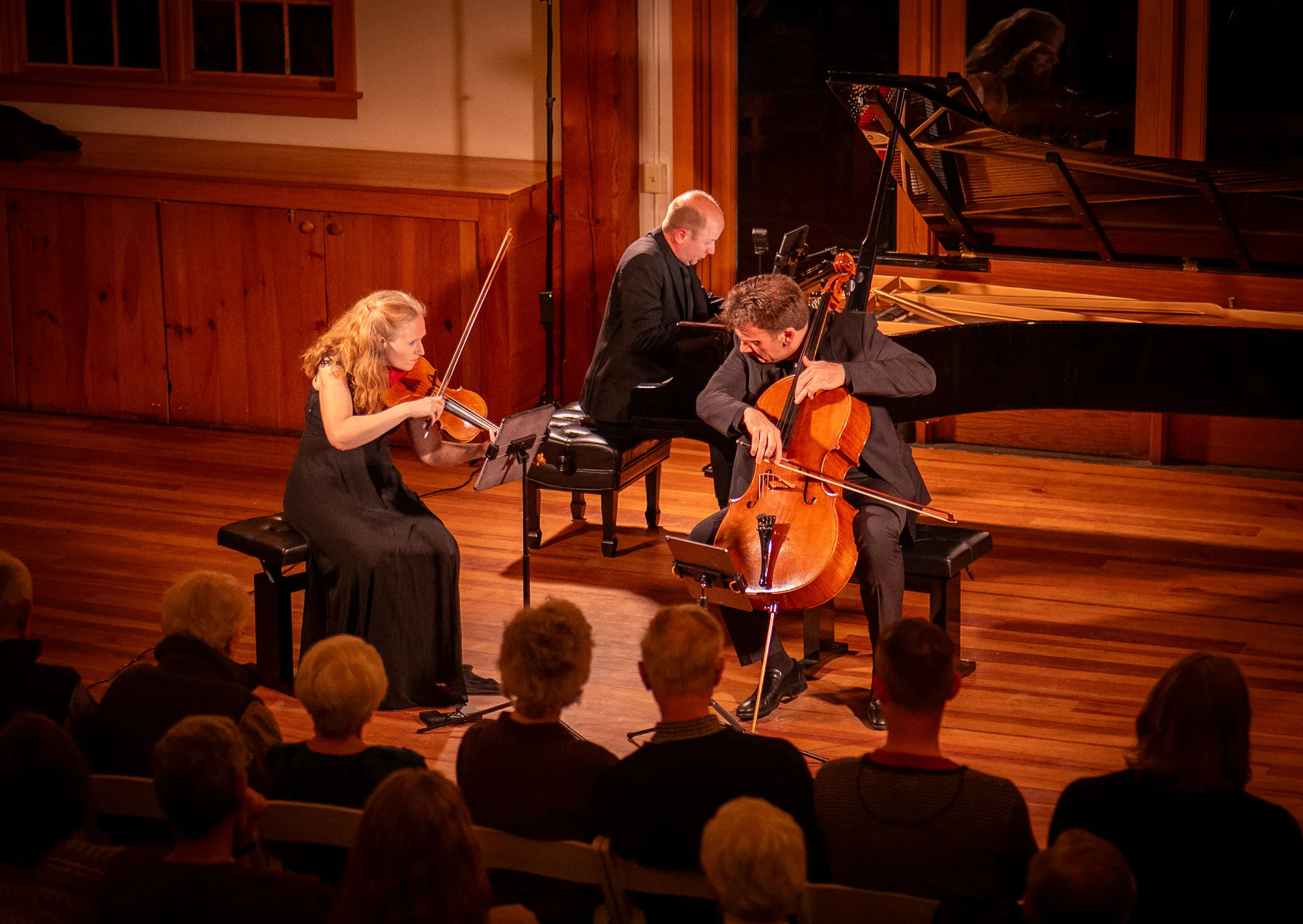 Three musicians perform on stage: a woman plays the violin, a man plays the cello, and another man plays the piano. They are in a warmly lit wooden room with an audience watching.