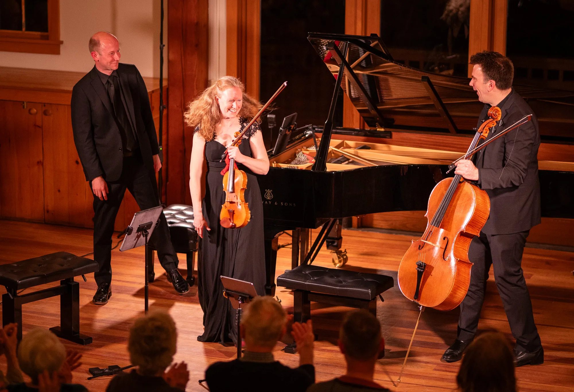 Three musicians onstage at a concert, smiling and holding their instruments—a violin, a cello, and a piano behind them—while the audience applauds. The setting is warmly lit with wooden floors and paneling.