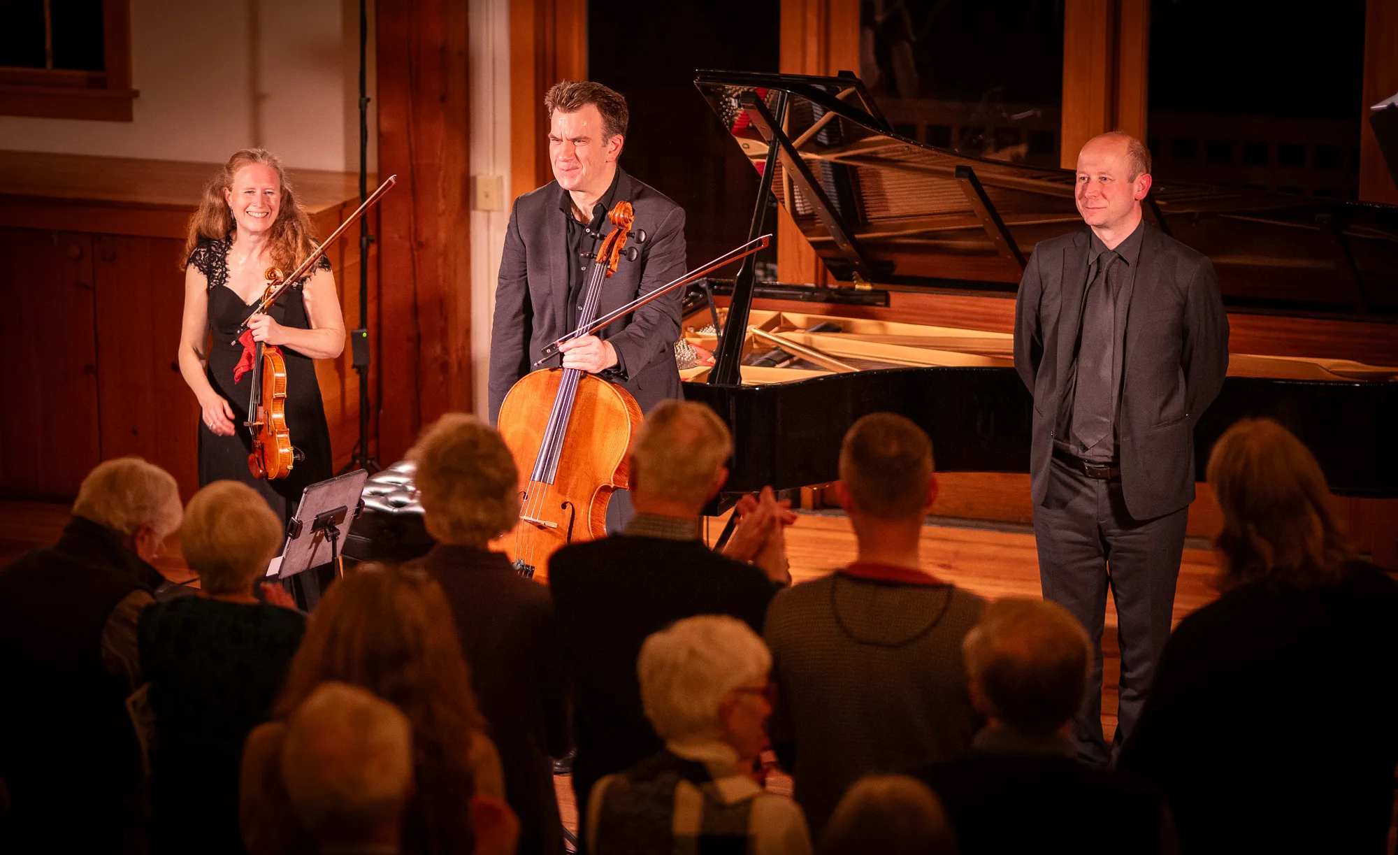 Three musicians—a violinist, cellist, and pianist—stand at the front of a stage, smiling and acknowledging applause from an audience in a warmly lit room with a grand piano in the background.