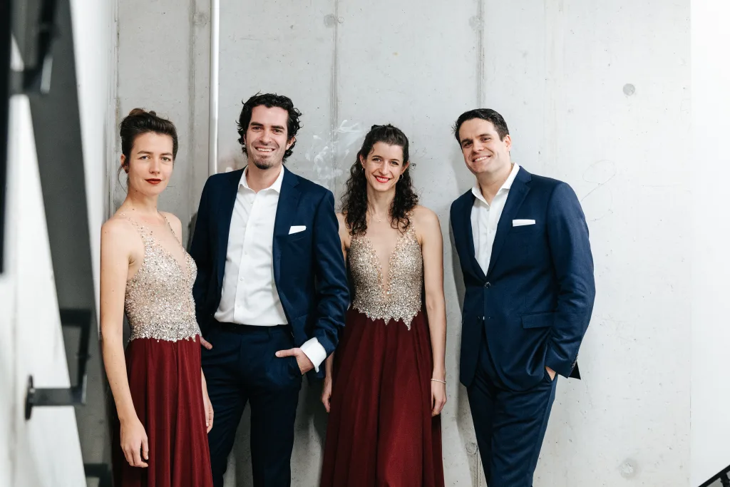 Four people from Ashuelot Concerts stand against a light-colored wall; two women wear glittery, sleeveless tops with long burgundy skirts, and two men wear dark blue suits with white shirts, all smiling and posing together.