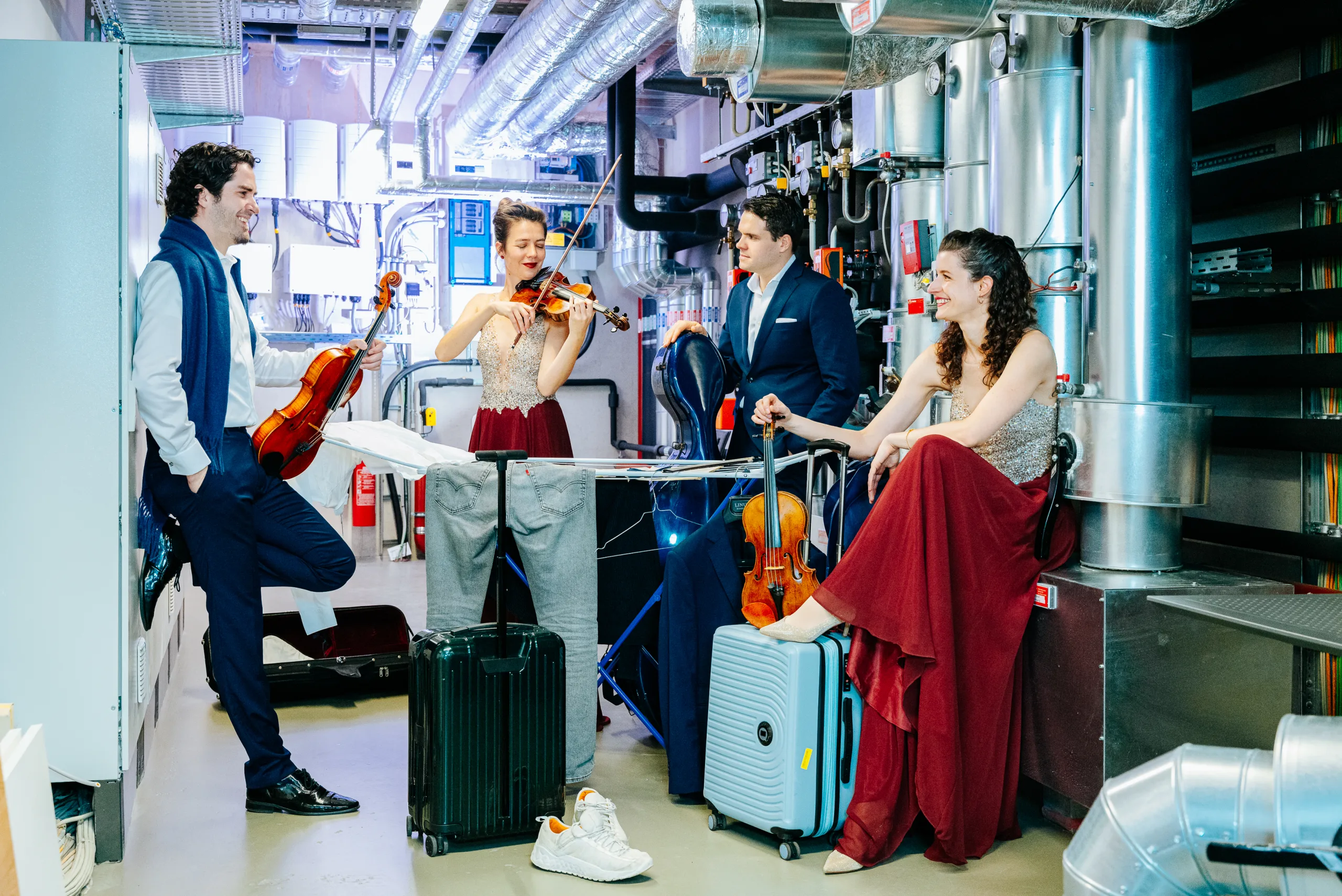 Four people in formal attire, with violins and a viola, stand and sit among luggage and pipes in an industrial backstage area, smiling and talking, as one person plays the violin.