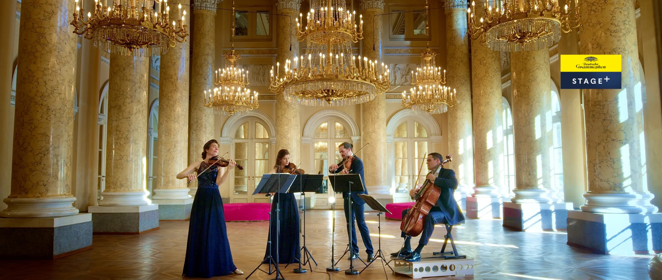 A string quartet performs in an opulent hall with marble columns, large chandeliers, and tall arched windows. Three musicians stand playing violins and viola, while one sits playing a cello.